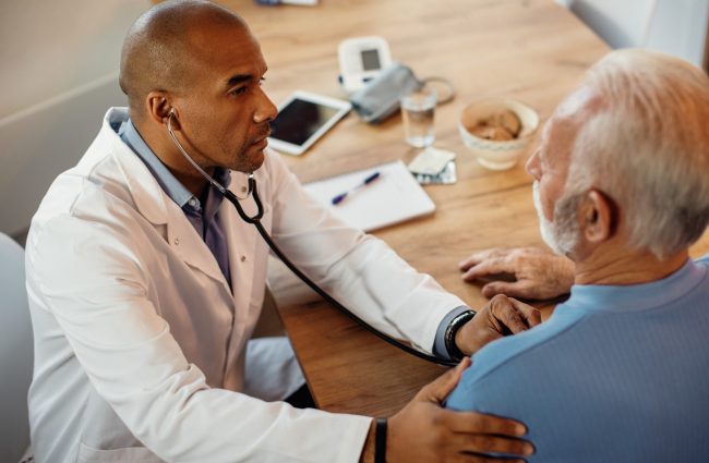 Black doctor examining senior patient with a stethoscope at nurs Photo of doctor with male senior patient illustrates blog "Does Medicare Pay for Nursing Home Care?"